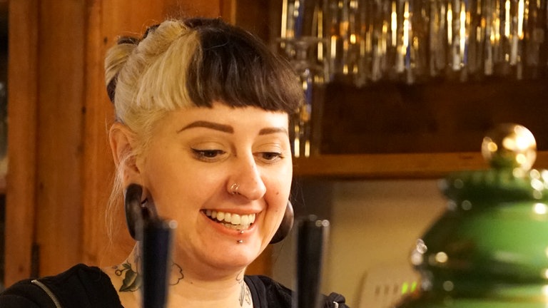 Junge Frau mit weiß-schwarzen Haaren steht grinsend am Bierzapfhahn und füllt ein Glas.