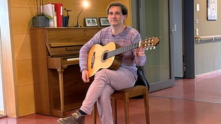 Ein Mann sitzt auf einem Stuhl und hat eine Gitarre in der Hand. Matthias begleitet als Musiktherapeut Menschen auf ihrem letzten Lebensweg.