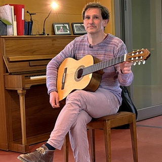 Ein Mann sitzt auf einem Stuhl und hat eine Gitarre in der Hand. Matthias begleitet als Musiktherapeut Menschen auf ihrem letzten Lebensweg.