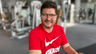 Mittelalter Mann mit braunen Haaren und Brille. Trägt ein rotes Shirt. Im Hintergrund sieht man Geräte aus einem Fitnessstudio.