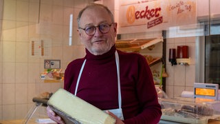 Ein Verkäufer hält einen Laib Käse in der Hand. Hanns wagt mit 63 einen Neuanfang und eröffnet einen Käseladen.