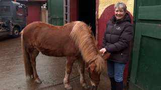 Eine Dame mit schwarzer Daunenjacke und kurzen Haaren steht vor einer offenen Stalltür. Neben ihr ein kleines braunes Pony.