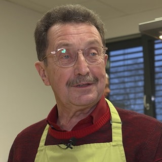 Älterer Herr mit braunen Haaren und Brille steht mit einer grünen Schürze und einem roten Strickpulli in einer Küche.