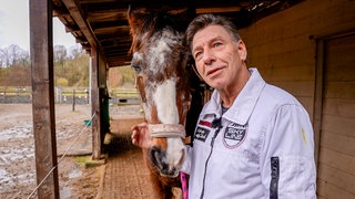 Mann mit grauen Haaren und weißer Jacke steht mit einem Pferd vor einem Pferdestall.