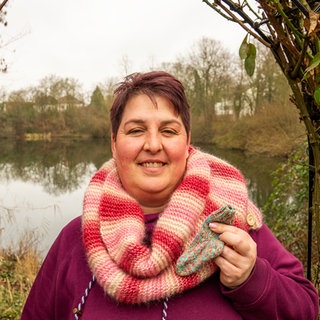 Frau mit roten Haaren trägt einen gestrickten Schal in pink und gelb. In der Hand hält sie ein kleines, bunt gestricktes Söckchen.