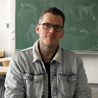 Junger Mann mit Brille, kurzen Haaren sitzt mit einer Jeansjacke bekleidet in einem leeren Klassenraum.