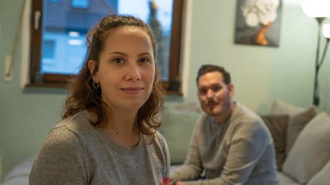 im Vordergrund sitzt eine junge Frau, Saskia, im Hintergrund ihr Ehemann, Sven
