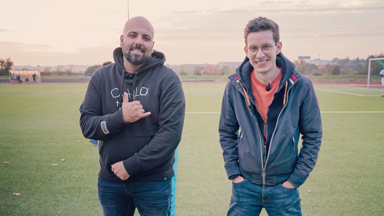 Zwei junge Männer stehen nebeneinander auf einem Fußballplatz.