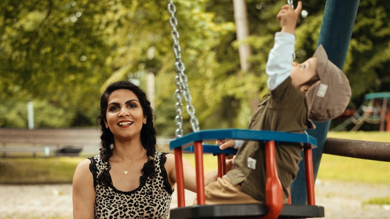 Frau mit ihrem Kind auf dem Spielplatz an einer Schaukel. 