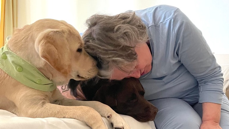 Hündinnen Sissi und Helga liegen auf einem Bett und eine alte Dame kuschelt mit ihnen.