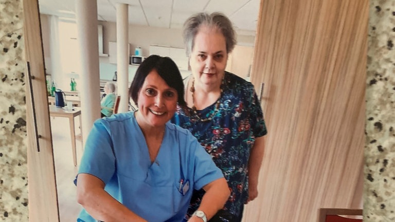 Frau in Pflegekleidung sitzt auf einem Rollator. Hinter ihr steht eine ältere Dame.