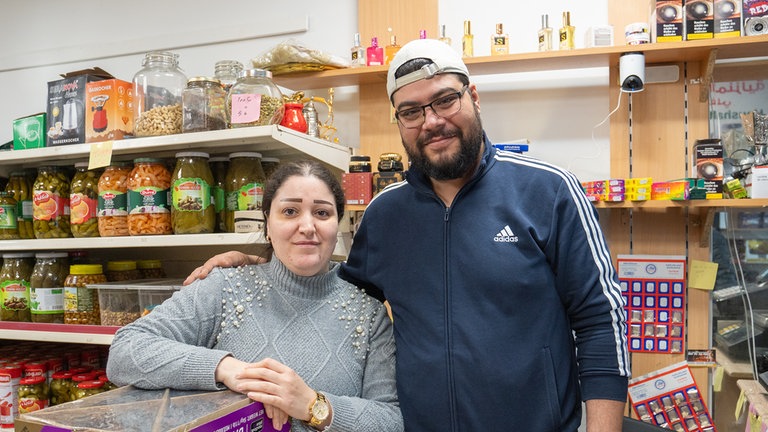 Ein Mann in Sportjacke und mit Mütze und eine Frau mit gruem Pullover schauen in die Kamera.