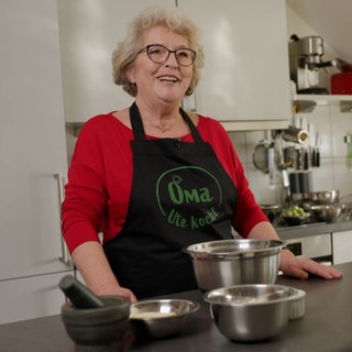 Frau mit weißen Haaren und schwarzer Schürze steht in Küche.