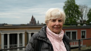 Ältere Frau mit weißen Haaren und rosa Pullover lächelt in Kamera.