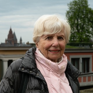 Ältere Frau mit weißen Haaren und rosa Pullover lächelt in Kamera.