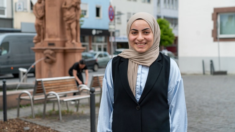 Eine junge Frau mit Kopftuch steht vor einem Brunnen und lächelt.