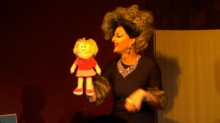 Eine Dragqueen steht mit einer Handpuppe auf einer Bühne
