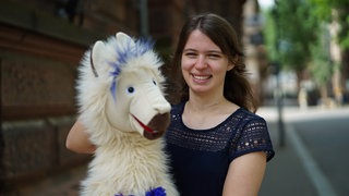 Frau mit braunen Haaren lächelt in Kamera, im Vordergrund unscharf eine Alpaka-Handpuppe.