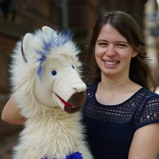 Frau mit braunen Haaren lächelt in Kamera, im Vordergrund unscharf eine Alpaka-Handpuppe.