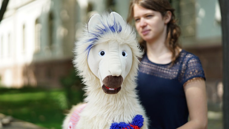 Ein Alpaka als Handpuppe im Vordergrund, unscharf dahinter eine Frau mit braunen Haaren.