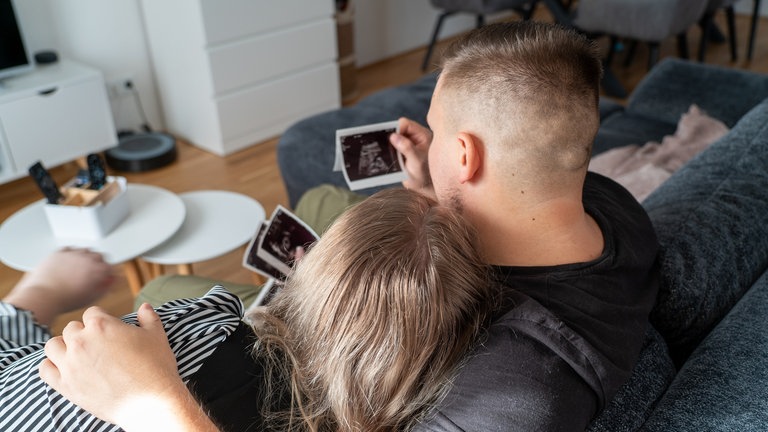 Junges Paar sitzt auf Sofa und schaut sich Ultraschallbilder an.