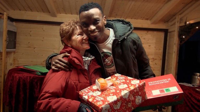 Ein junger schwarzer Mann umarmt eine ältere Frau, die einen Schuhkarton hält.