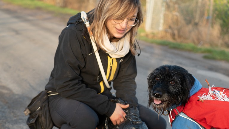 Hund schaut in die Kamera und hockt neben blonder Frau mit Brille.
