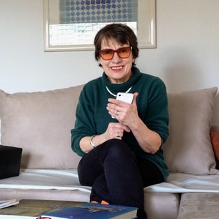 Frau mit braunen Haaren, getönter Brille und grünem Pullover lächelt in Kamera.