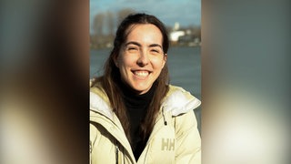 Junge Frau mit dunklen Haaren und beiger Jacke lächelt in Kamera.