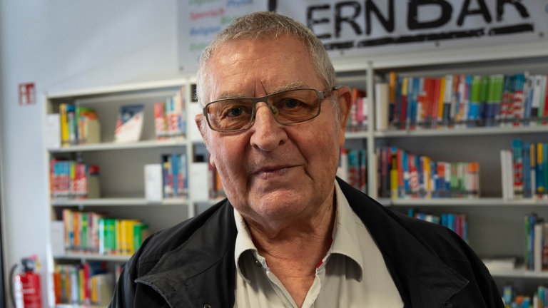 Ein älterer Mann, mit kurzen Haaren und Brille. Er trägt ein helles Hemd und darauf eine dunkle Jacke. 