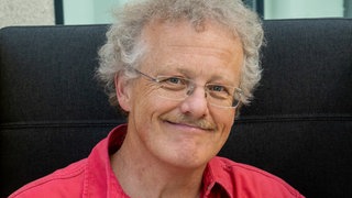 Älterer Herr mit rotem Hemd und Brille lacht in die Kamera.