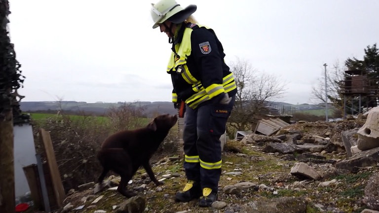 Eine Frau in Feuerwehrkleidung beugt sich zu ihrem dunkelbraunen Hund. Hinter ihnen befindet sich Schutt.