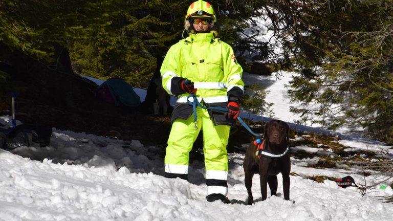 Frau trägt einen leuchtgelben Anzug mit Reflektoren und einen gelben Helm. Mit der linken Hand hält sie ihren dunkelbraunen Hund an der Leine fest. Beide stehen im Schnee. 
