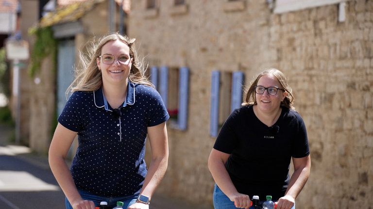 Zwei junge Frauen fahren mit dem Tretroller durchs Dorf.