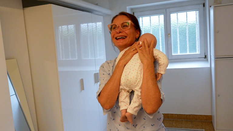 Eine Frau mit dunklen Haaren, Zopf und Brille schaukelt lachend eine Babypuppe.