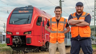 Zwei junge Männer mit Warnwesten posieren vor einer Lok