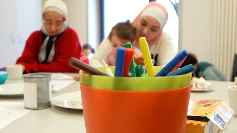 Im Vordergrund steht ein Becher mit bunten Stiften, im Hintergrund sitzen zwei Frauen mit Kopftuch, eine von ihnen hat ein Kind auf dem Schoß.