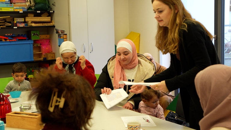 Frauen mit Kopftuch sitzen um einen Tisch, eine Frau verteilt Blätter