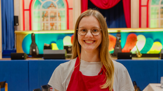 Junge blonde Frau mit Brille steht vor bunter Bühne und lächelt mit roter Schürze und weißem T-Shirt.