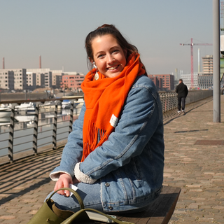 Eine junge Frau, sitzt lächelnd auf einer Bank. Hat braune Haare, trägt eine Jeansjacke und einen knall orangenen Schal.
