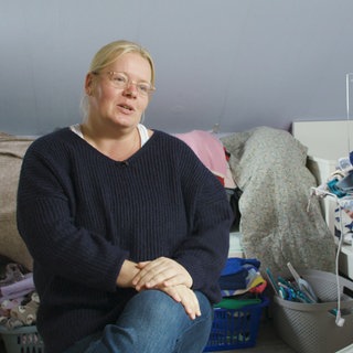 Eine blonde Frau mit Brille sitzt vor Wäschebergen.  