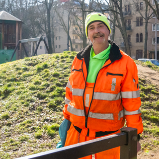Mitarbeiter der Müllabfuhr. Trägt einen orangenen Anzug und eine grüne Mütze.