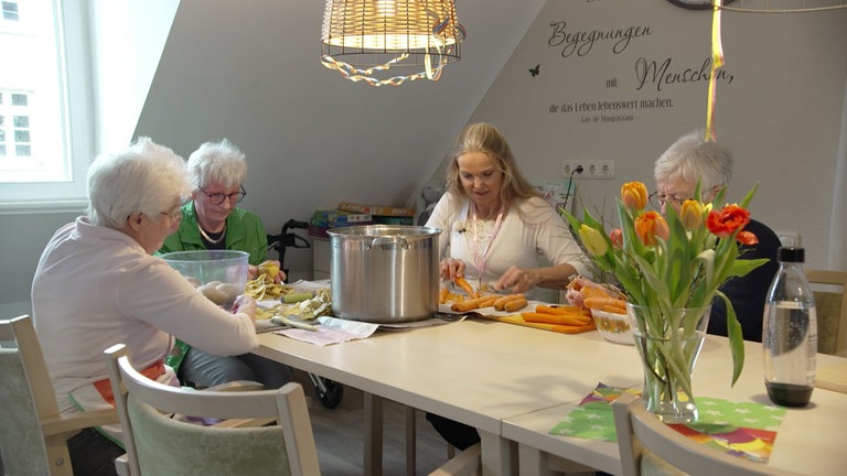 Zwei ältere Frauen und eine junge Frau beim Kartoffeln und Karotten schälen.