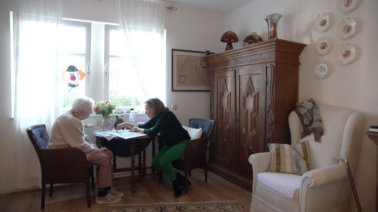 Eine ältere und eine jüngere Frau sitzen gemeinsam am Tisch und schauen sich Fotoalben an.