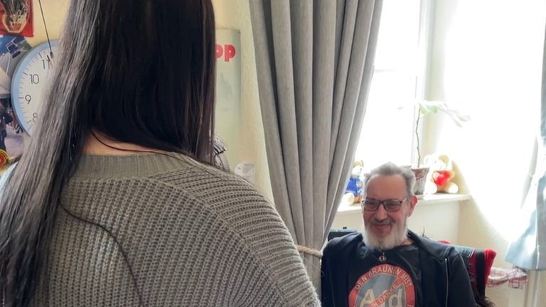 Ein älterer Mann lächelt eine junge Frau an, die mit dem Rücken zur Kamera steht.