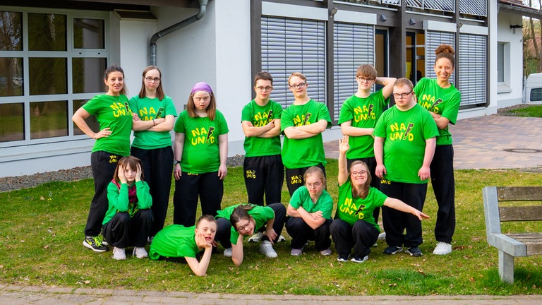 Eine Gruppe Jugendlicher mit Downsyndrom steht mit zwei erwachsenen Frauen auf einer Wiese und lächeln. Alle tragen grüne T-Shirts.