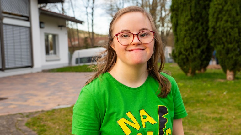 Ein Mädchen mit blonden Haaren und Downsyndrom lächelt in die Kamera. Sie trägt ein grünes T-Shirt und eine Brille.