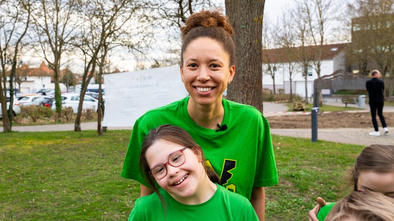 Eine Frau mit braunen Haaren und grünem T-Shirt lächelt in Kamera, vor ihr ein Mädchen mit Downsyndrom und grünem T-Shirt, das lächelt.