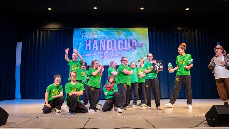 Mehrere Jugendliche mit Downsyndrom stehen in grünen T-Shirts auf einer Bühne mit zwei Erwachsenen. 