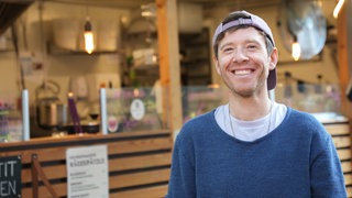 Mann in blauem Pullover mit Kappe lächelt in die Kamera.